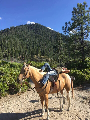 Cindy Hartzell lays across the back of a palomino horse near some woods with a mountain in the backdrop.